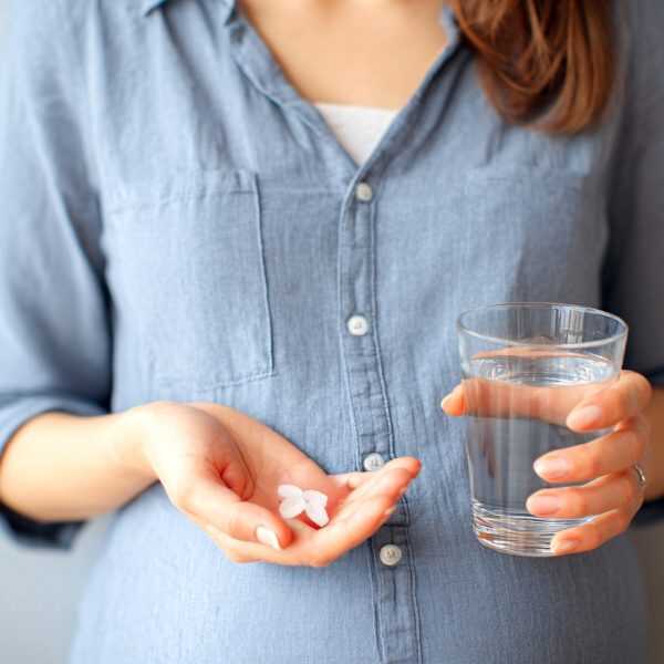 A woman in a denim shirt holds a glass of water in one hand and several white pills in her other open palm.