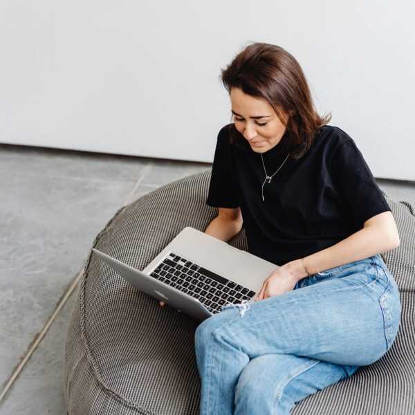 A woman in a black t-shirt and blue jeans sits in a grey beanbag chair, looking down at a silver laptop on her lap in a minimalist room.